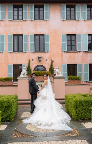 A wedding couple stands in front of a pink Mediterranean-style villa with pale blue shutters and manicured hedges.