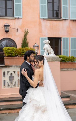 Newlyweds embrace on steps of peach building with white statue and blue shutters in romantic outdoor setting.