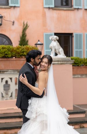 Wedding couple shares intimate moment by peach colored villa with dog statue and greenery.