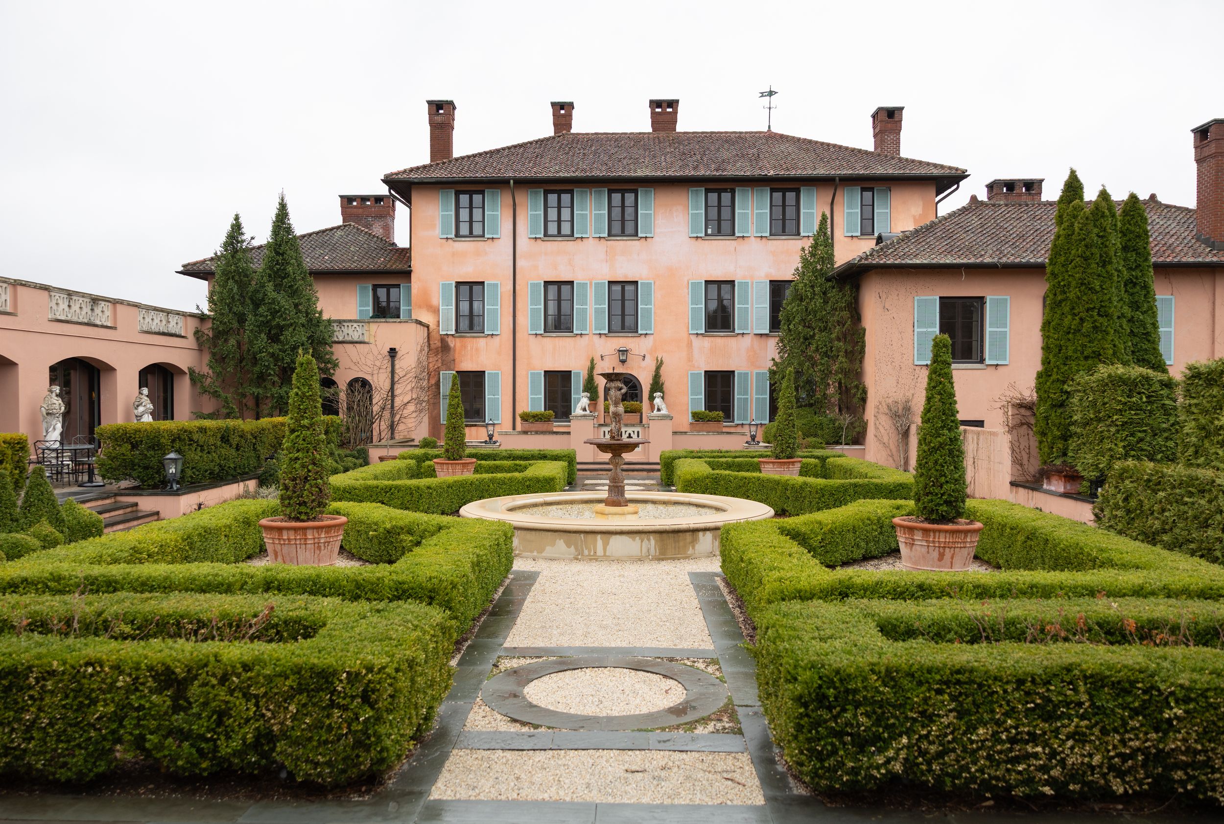 Italian villa with formal gardens featuring symmetrical hedges and central fountain surrounded by cypress trees.