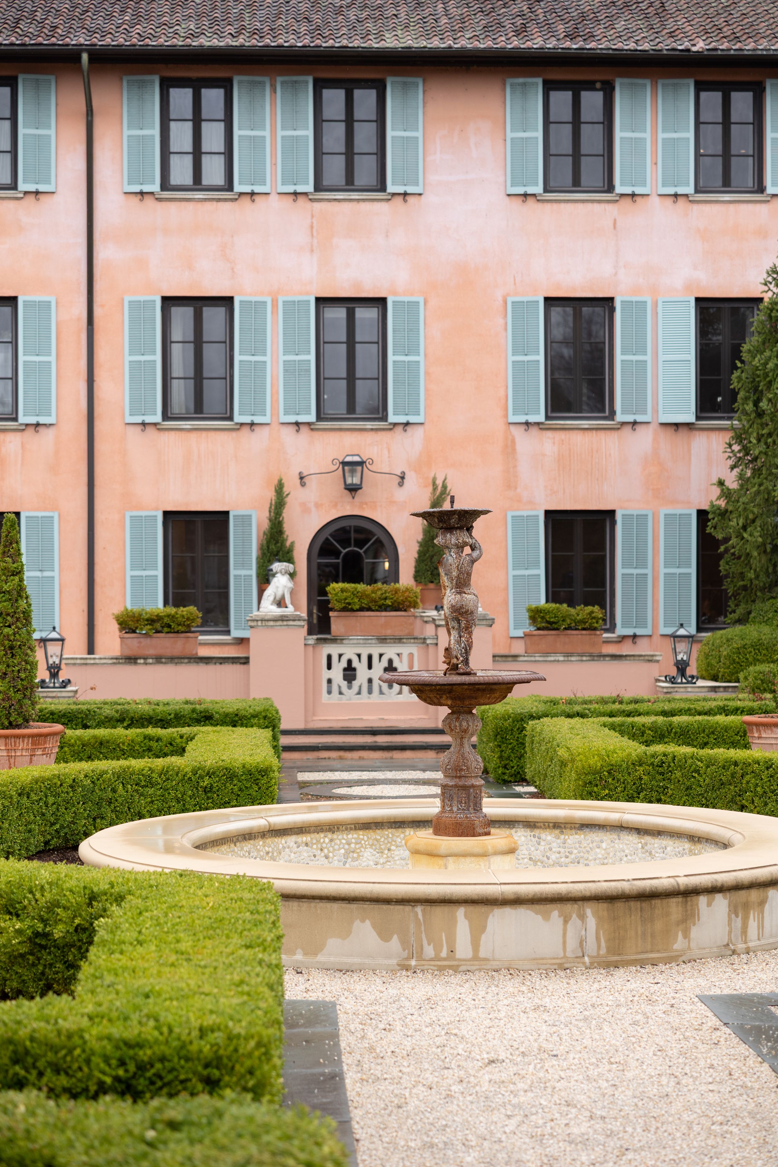A peachy pink villa with blue shutters and a formal garden featuring a classical fountain surrounded by manicured hedges.