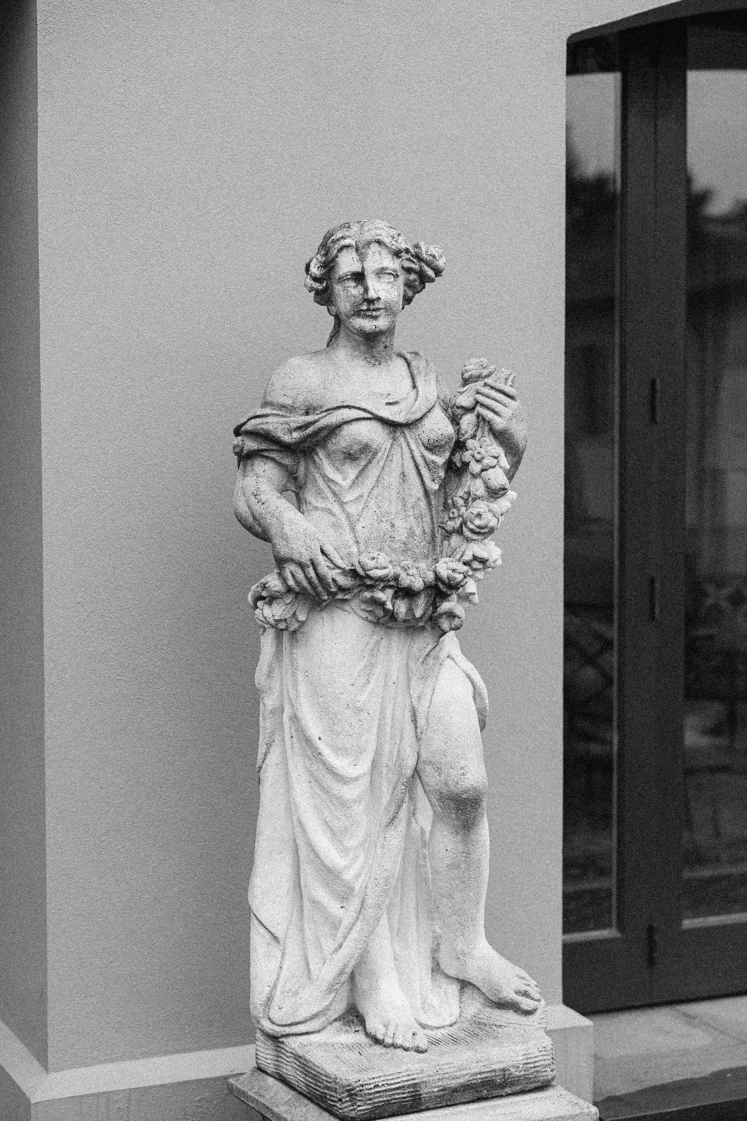 Classical stone statue of a woman in draped clothing holding flowers in a garden setting.