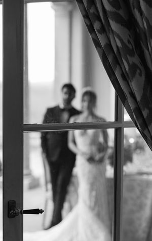 A black and white silhouette of a wedding couple viewed through a window with a curtain.