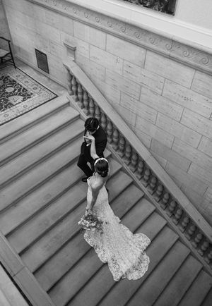A couple descending a grand marble staircase in formal wedding attire, captured in black and white.