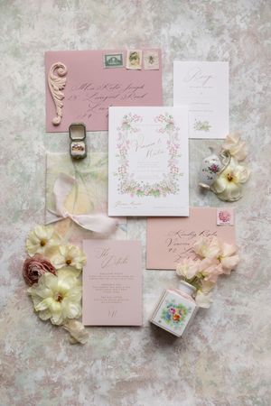 Elegant wedding invitation suite in blush pink with floral details, rings, and delicate flowers arranged on textured surface.