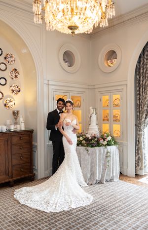 Wedding couple poses in elegant room with crystal chandelier and wedding cake display.