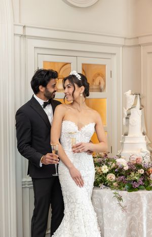 An elegant wedding couple sharing an intimate moment beside a white wedding cake and floral arrangements.