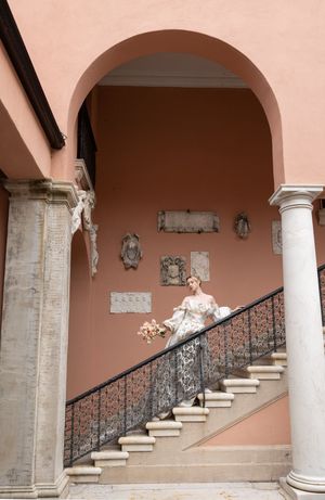 A bride in white gown ascending a classical arched staircase with pink walls and ornate columns.