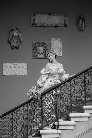 Black and white photo of a bride in an elegant gown posing on an ornate staircase with wall decorations.