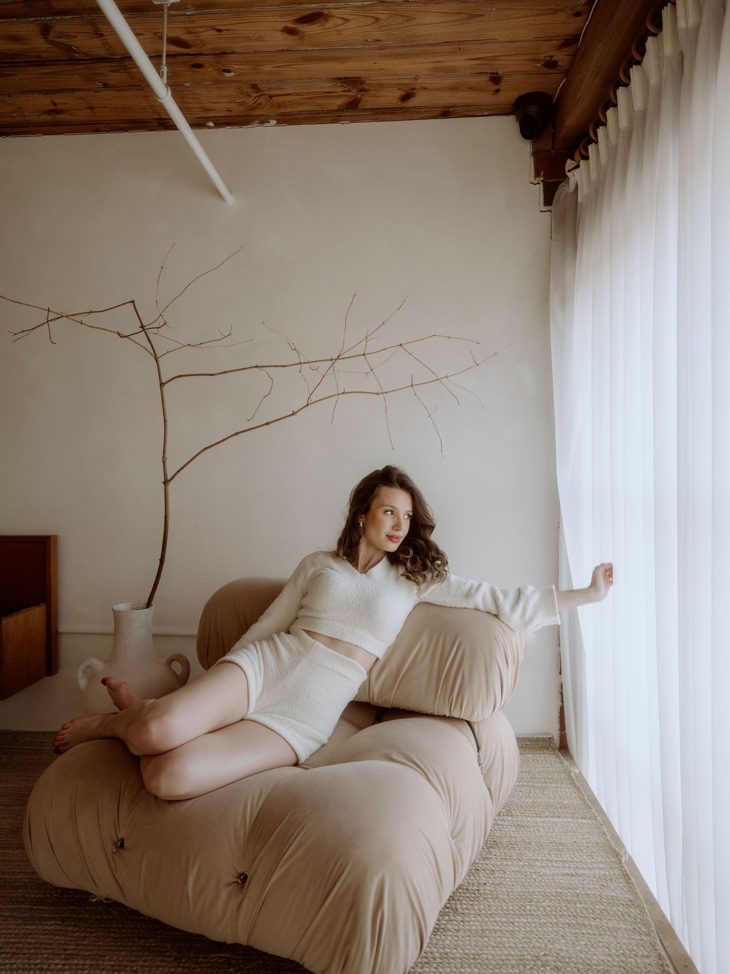 A person reclines gracefully on a beige lounge chair near a window with natural light and minimal decor.