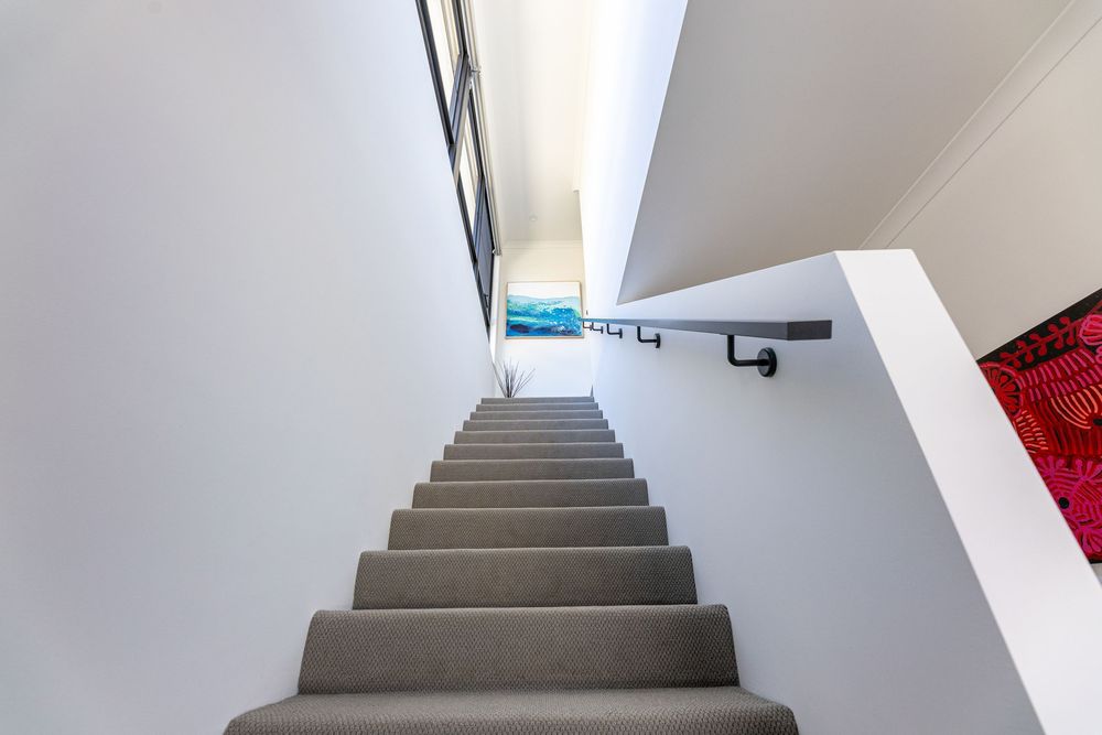 Modern staircase with grey carpeting leading upwards between white walls with a black handrail and bright natural light.