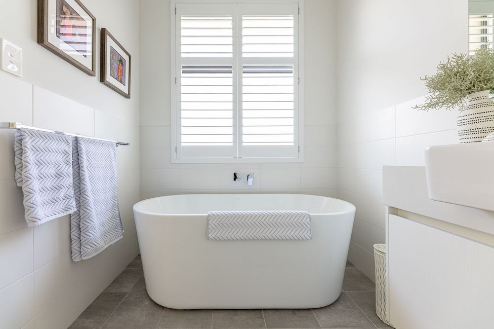 A classic white freestanding bathtub in a bright, minimalist bathroom with gorgeous plantation shutters and grey tile flooring.