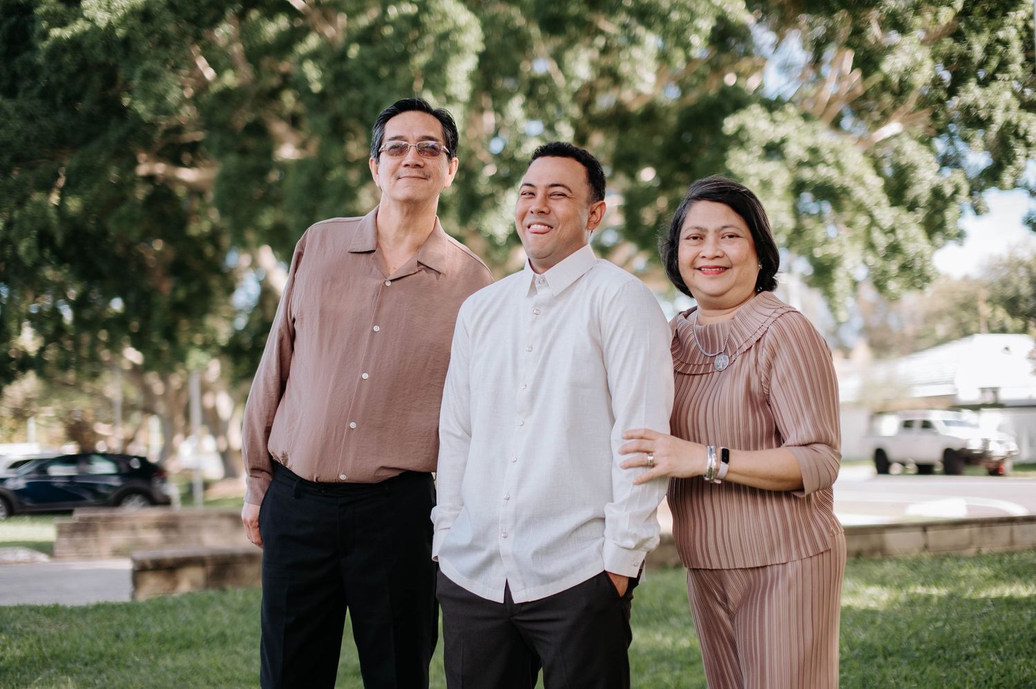 Three people stand together smiling outdoors in coordinated casual outfits against a sunlit garden background.