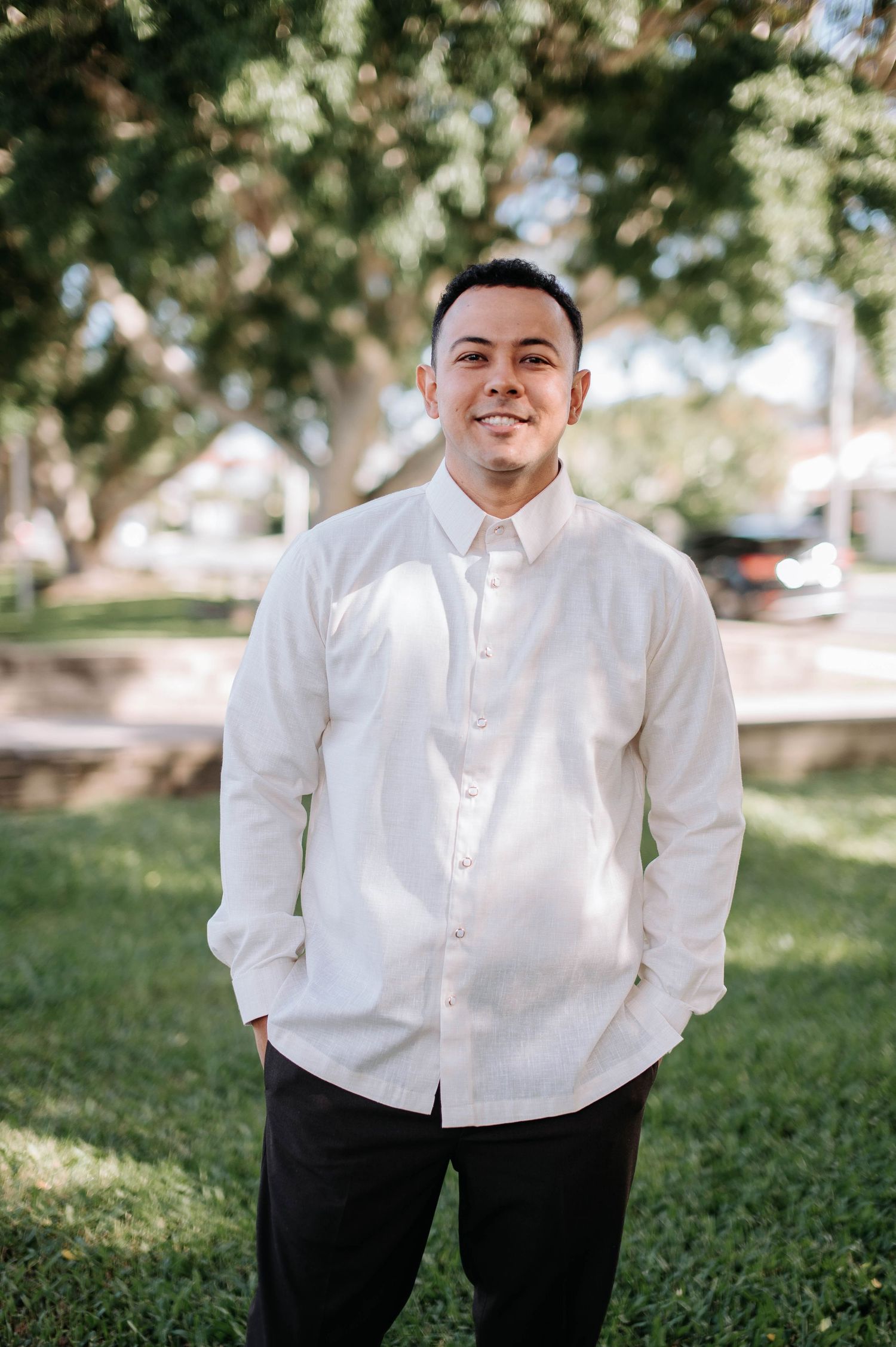Person in white button-down shirt and black pants standing outdoors in a park setting with trees in background.