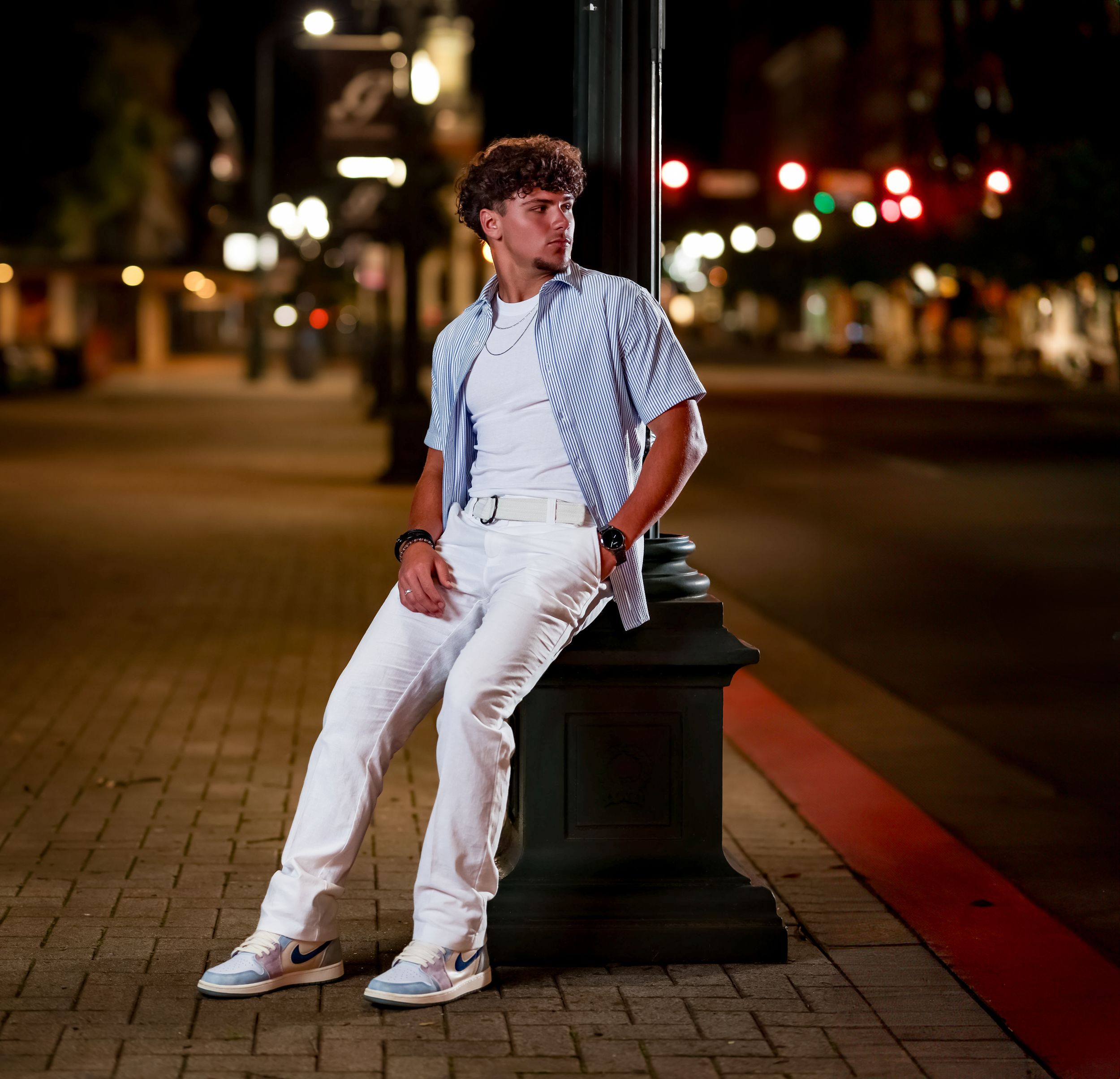 Person in light blue shirt and white pants leaning against lamp post at night with city lights in background.