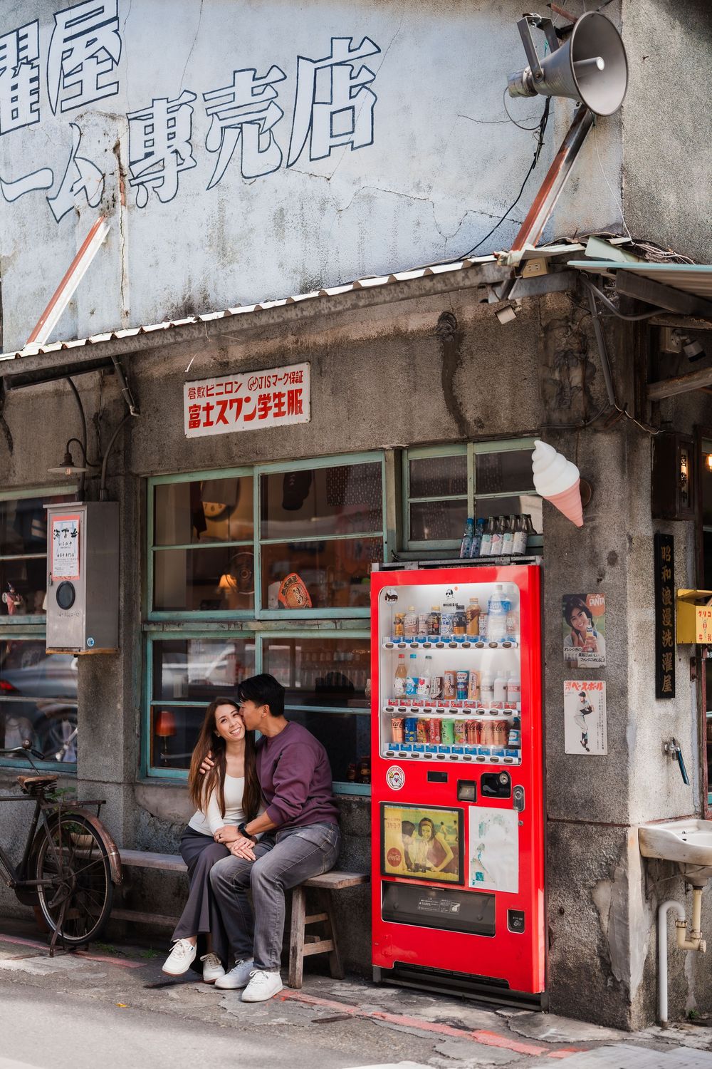 A couple sits outside a small Japanese shop with a red vending machine and vintage storefront at Dihua Street of Dadaocheng.