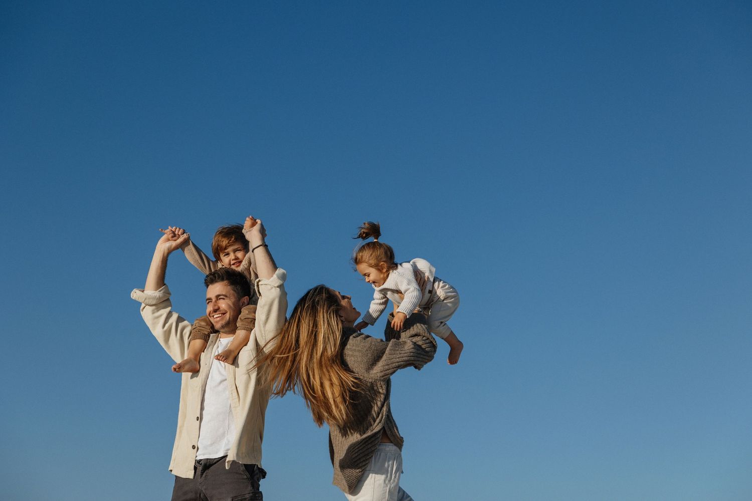 Happy family with two young children on shoulders playing under clear blue sky outdoors.