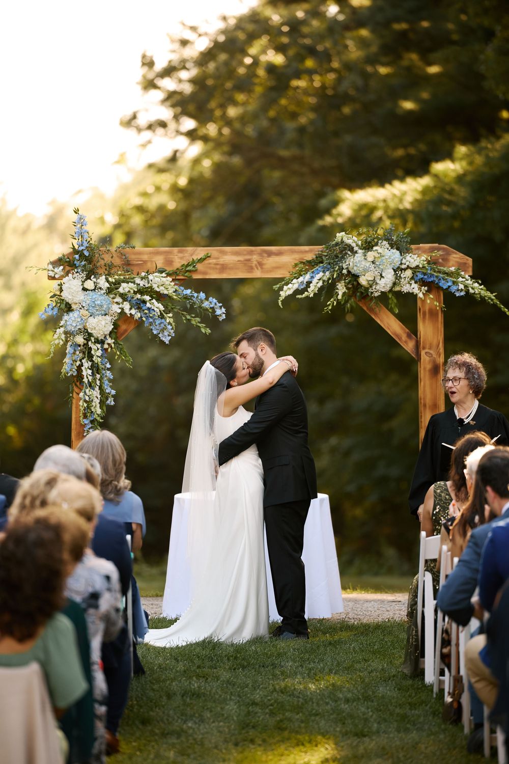Bride and groom share first kiss under rustic wooden floral arch at outdoor wedding ceremony with seated guests.