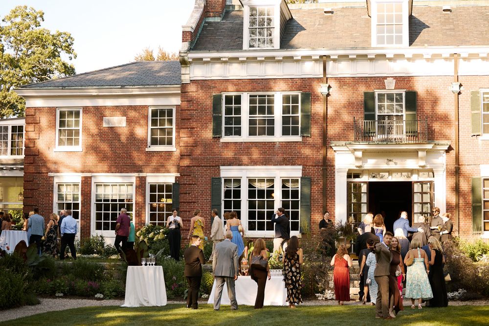 Elegant outdoor garden party at a brick colonial mansion with guests mingling on the lawn at dusk.