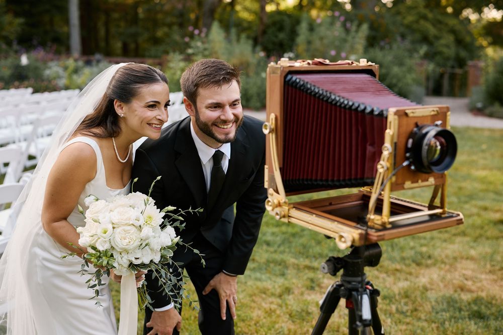 Bride and groom smiling beside a vintage large format wooden camera on a tripod at an outdoor wedding venue.