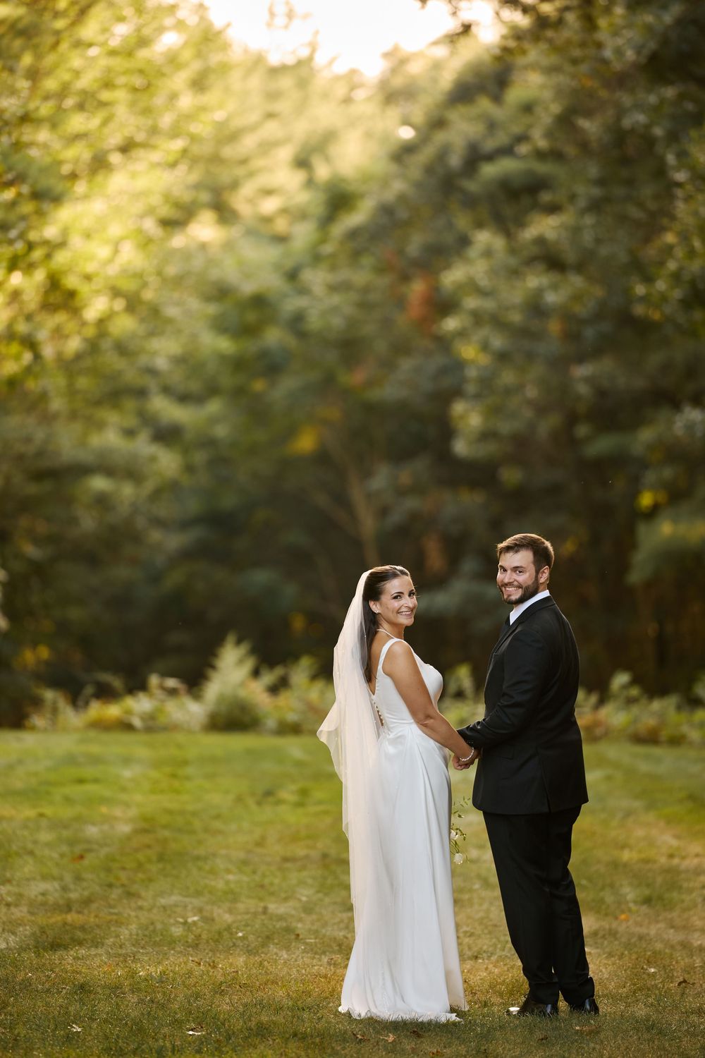 Bride and groom holding hands on green lawn with lush forest backdrop on their wedding day.
