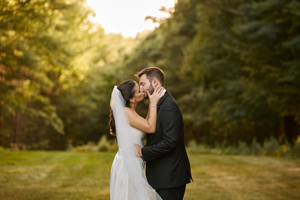 Bride and groom share a romantic kiss outdoors at golden hour, surrounded by lush green trees.