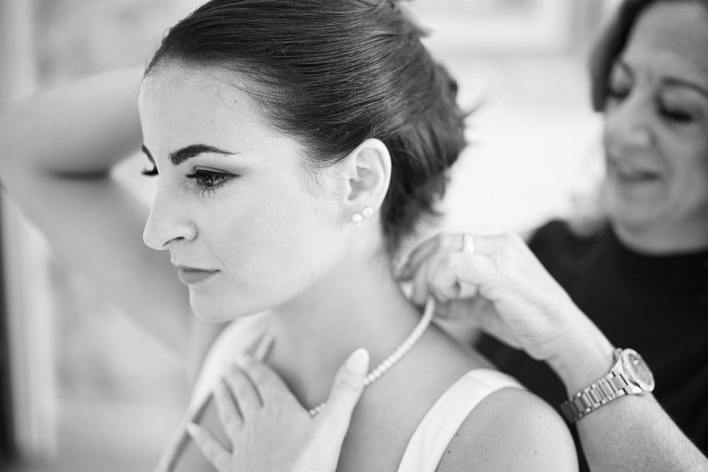 Black and white photo of a bride having pearl necklace fastened by bridesmaid during wedding preparations.