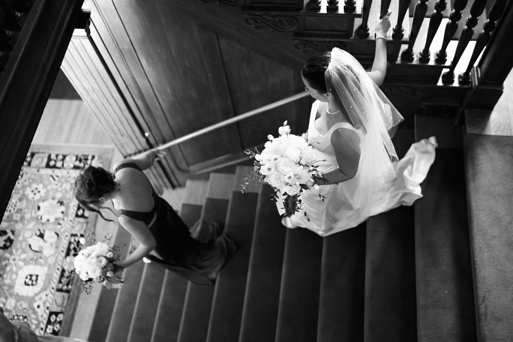 Black and white wedding photo of bride holding bouquet descending grand staircase with groom waiting below.