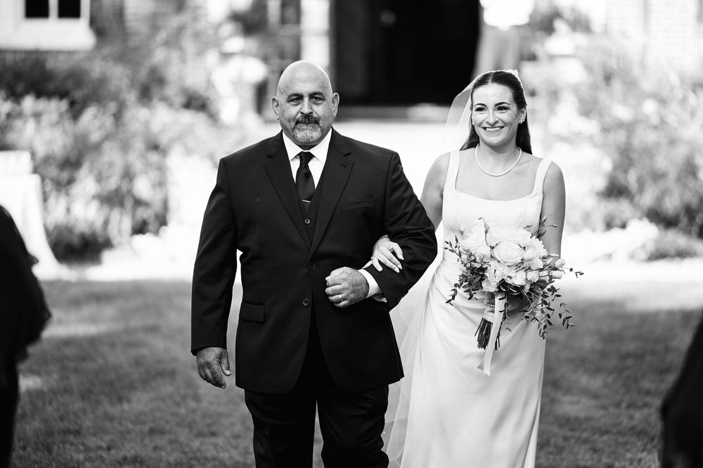 Black and white photo of a bride holding a bouquet walking down the aisle with an older man at an outdoor wedding.