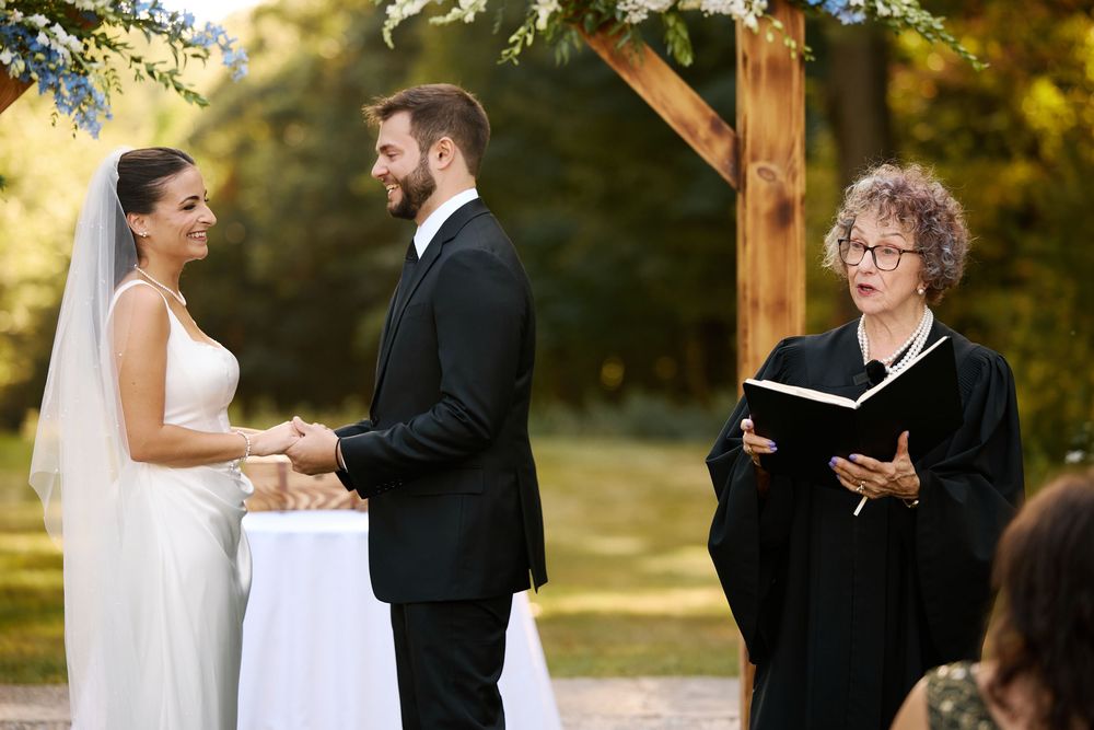 Couple holds hands during outdoor wedding ceremony while officiant reads from book under wooden arch.