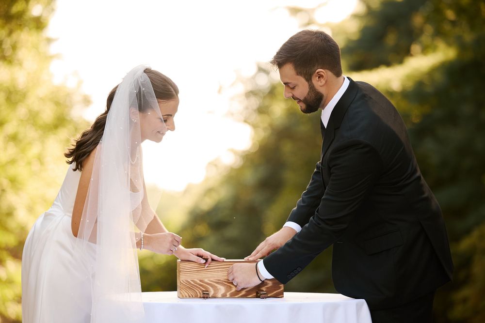 Bride and groom signing wedding documents at outdoor ceremony table, surrounded by lush greenery in golden sunlight.