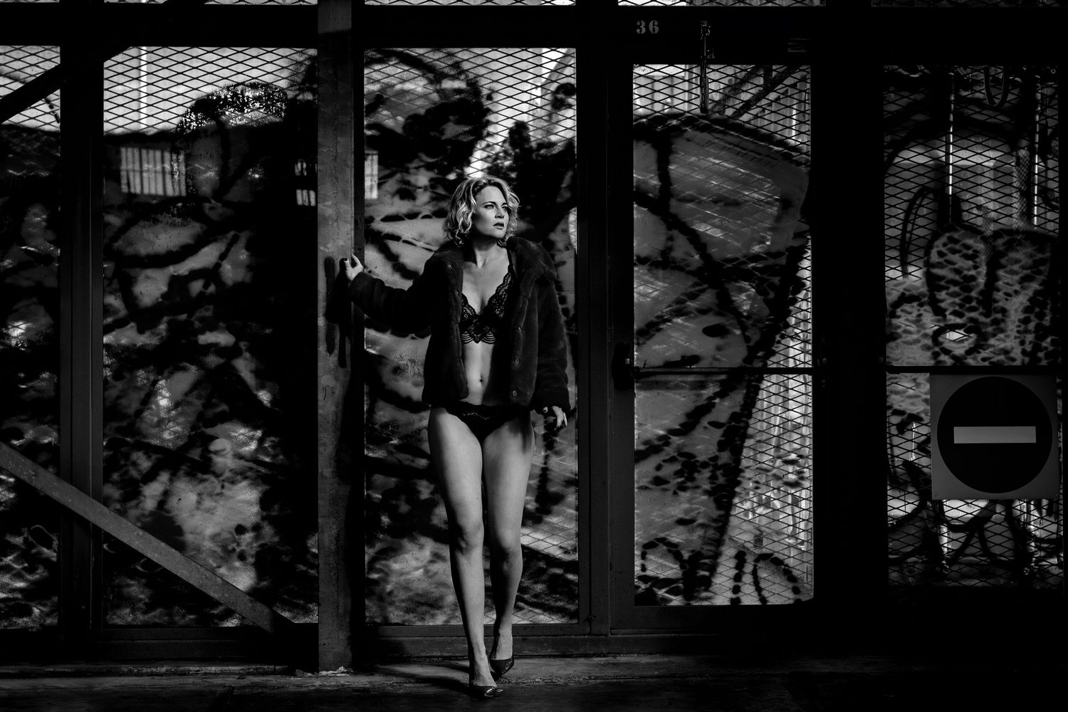 Black and white boudoir photo of a woman in lace lingerie posing by a window with dramatic shadows.