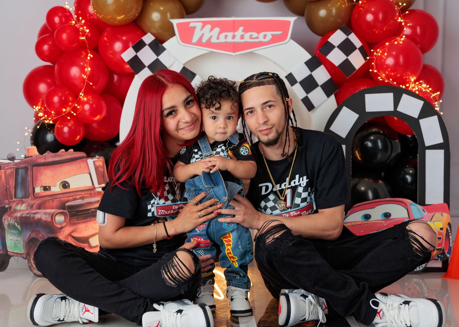 Family celebrating baby Matco's first birthday with a Cars-themed party featuring red balloons and checkered decorations.