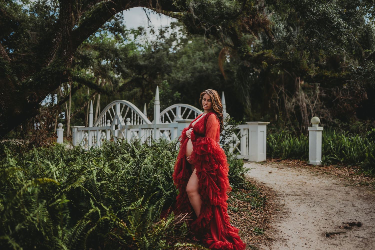 Pregnant woman in flowing red gown poses gracefully in lush garden with white gate and ancient trees.