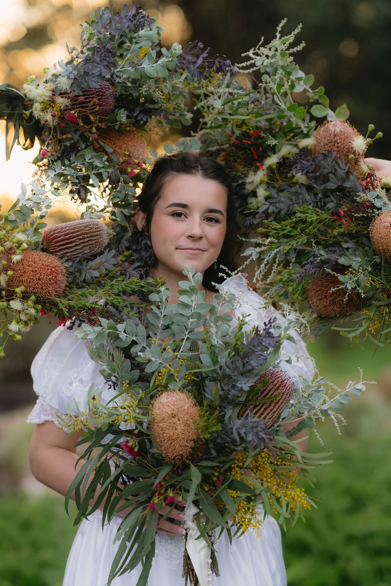 A tattooed woman raises a vibrant wedding bouquet of purple alliums, orange and red roses, and green foliage outdoors.