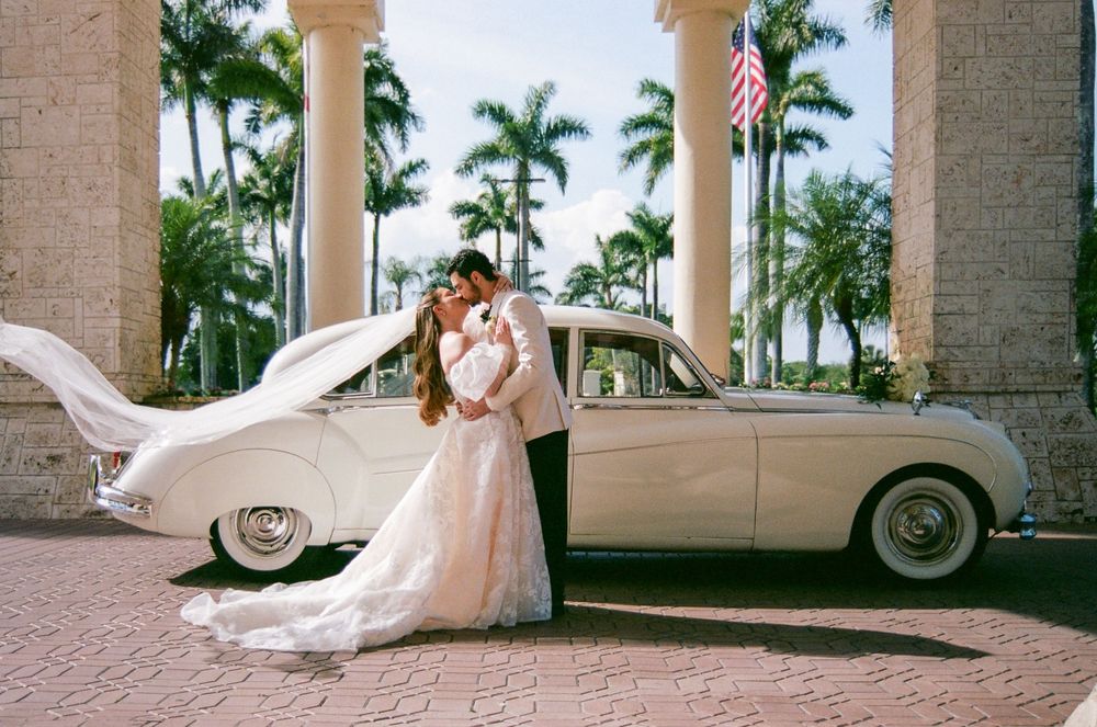 Bride and groom kissing beside a vintage white Rolls-Royce at a tropical wedding venue with palm trees.