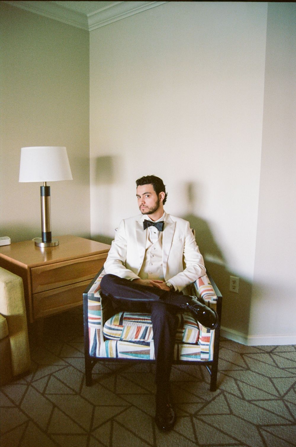 Groom in white tuxedo jacket and bow tie sits in a colorful chair in a hotel room.