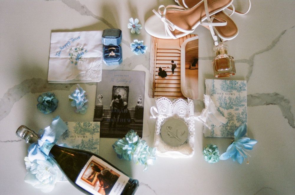 Elegant wedding flat lay with blue toile details, vintage photos, champagne bottle, heels, and floral accents on marble.