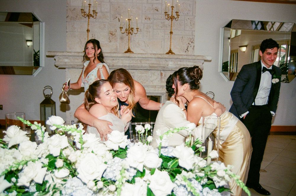Bridesmaids embracing emotionally at wedding reception table adorned with white roses and greenery floral arrangements.