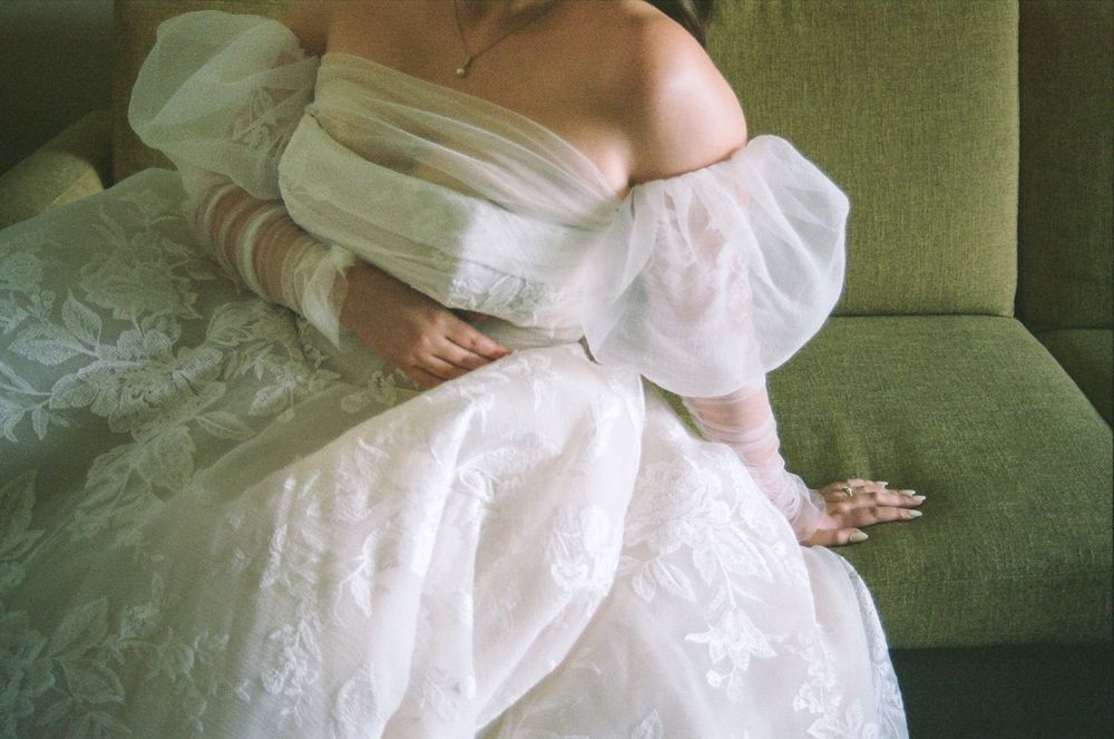 A bride in an elegant off-shoulder white lace wedding gown with puff sleeves sits gracefully on a green velvet sofa.