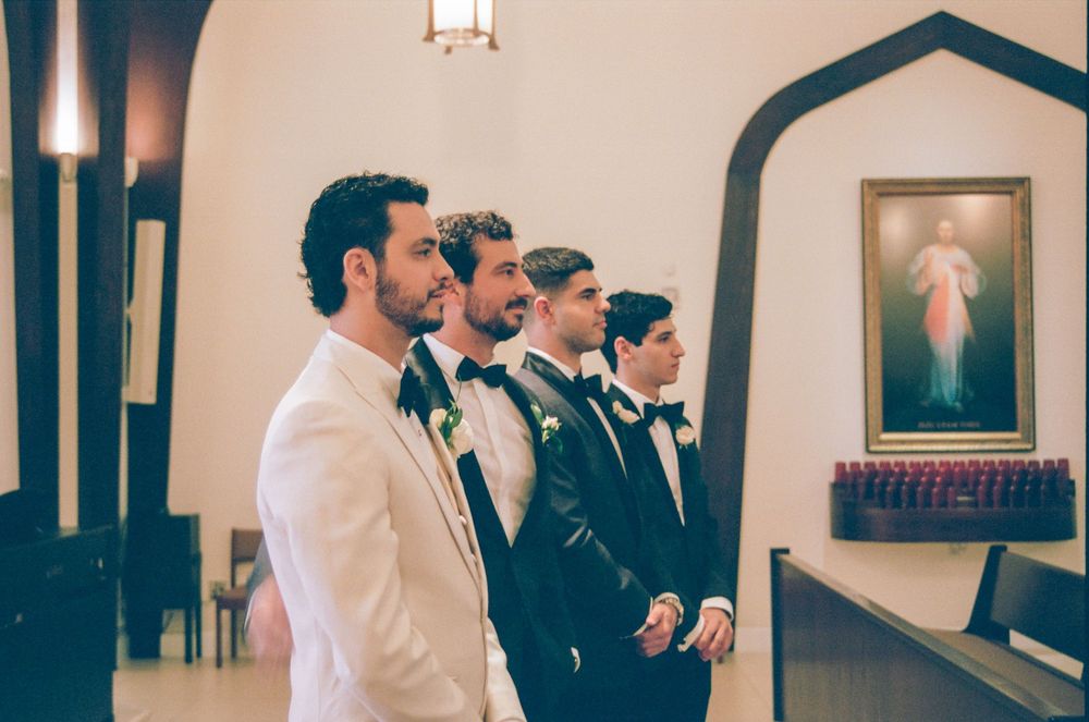 Groomsmen in tuxedos stand at church altar during wedding ceremony with religious painting on wall.