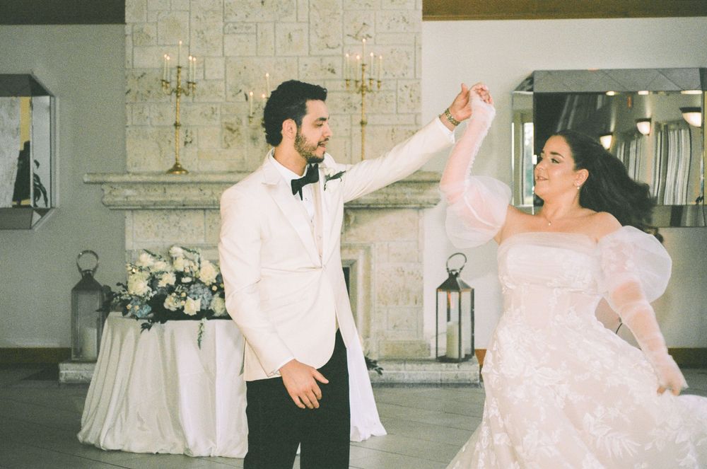 Bride and groom share first dance, groom in white tuxedo twirling bride in fluffy white gown at elegant wedding reception.