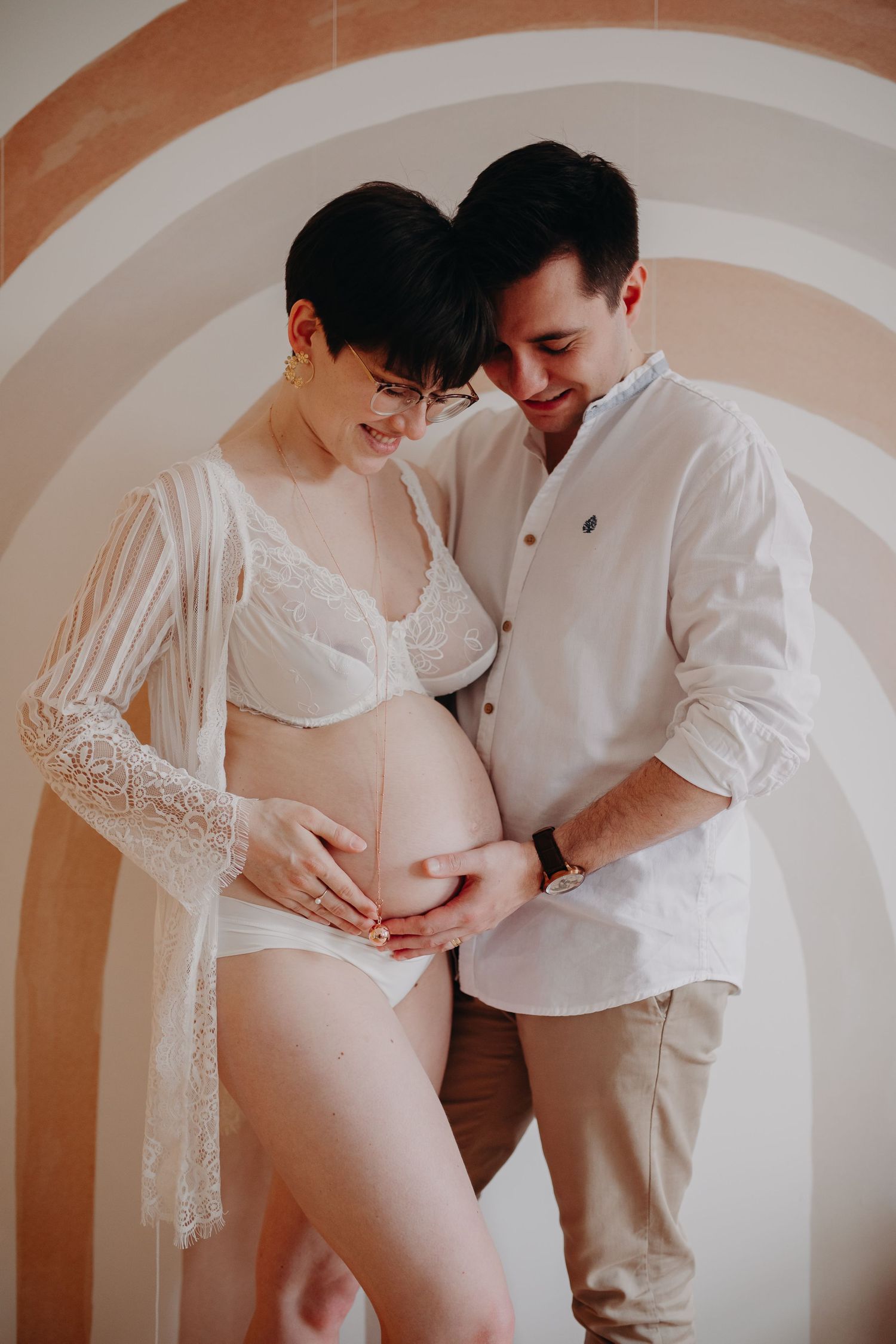 Smiling pregnant couple in white outfits embracing in front of a rainbow wall mural.