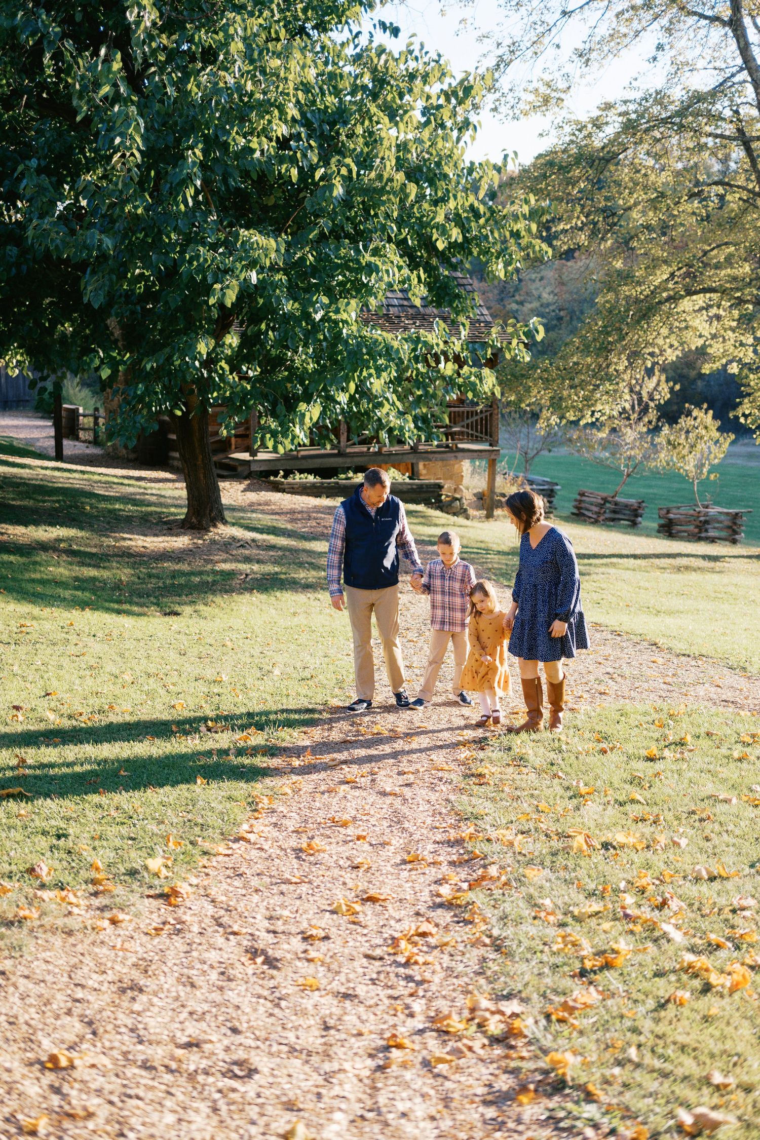 Family of four walking hand-in-hand on a sunny autumn park path surrounded by green trees and fallen leaves.