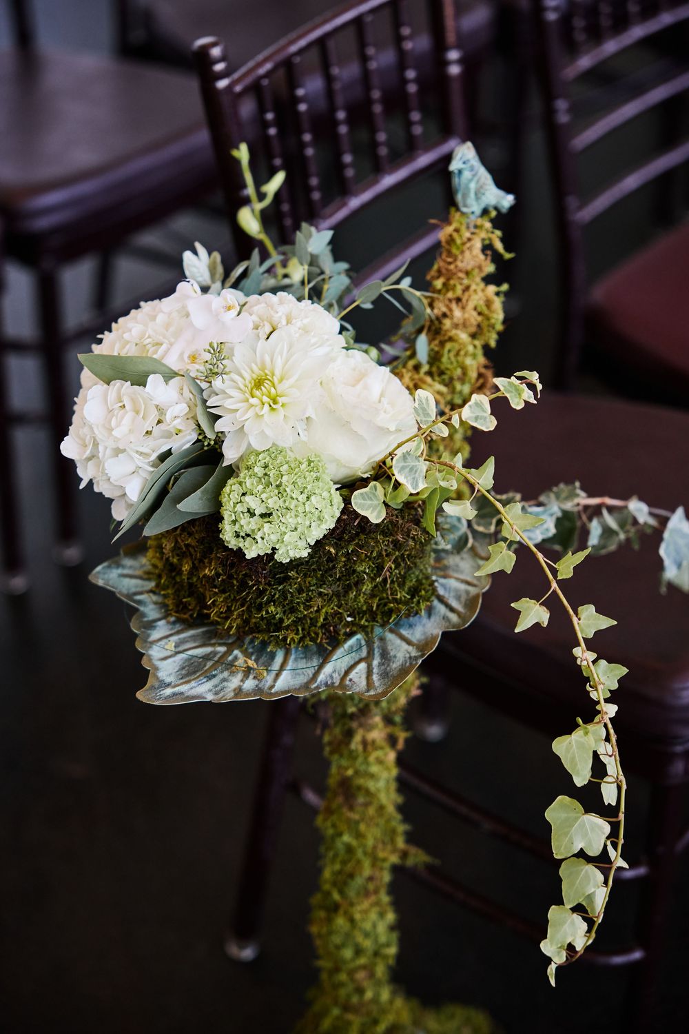 White roses, hydrangeas, and ivy pew floral arrangement on moss-covered stand at wedding ceremony.