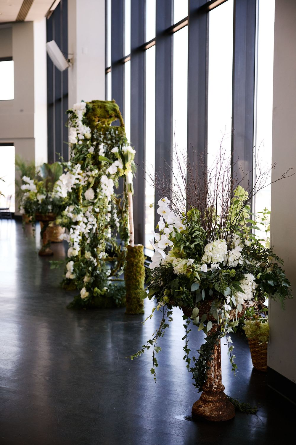 Elegant white floral arrangements with greenery line a modern wedding venue with floor-to-ceiling windows.