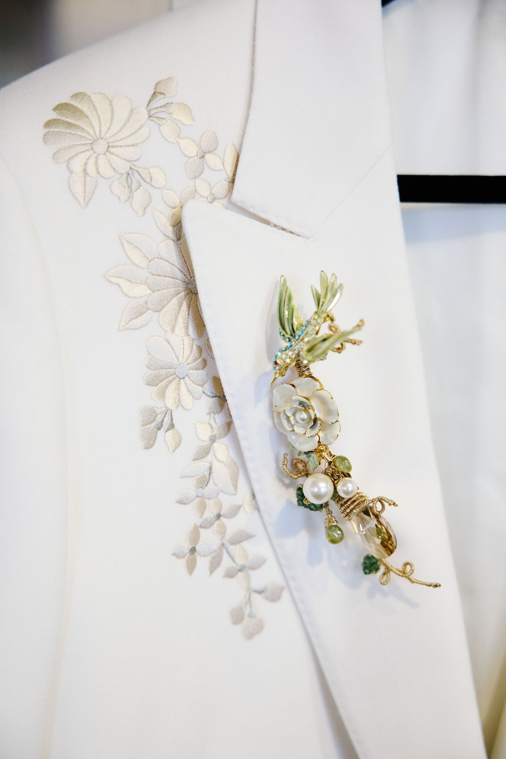 Close-up of an ivory embroidered wedding tuxedo lapel adorned with an elegant gold floral brooch and pearl accents.