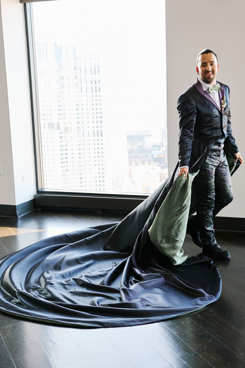 Groom in black tuxedo with dramatic long dark train poses by floor-to-ceiling window with city skyline views.