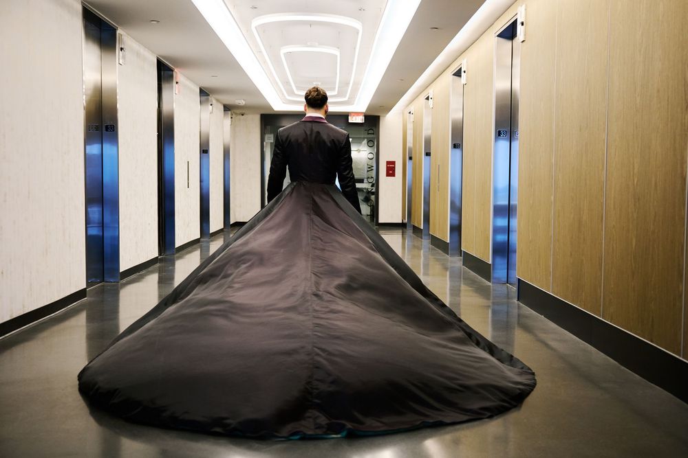 Person in a black suit with an extraordinarily long dramatic train filling a modern hotel corridor with elevator doors.