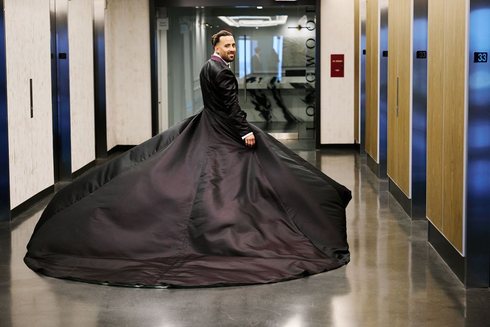 Groom in black wedding gown with long train twirls in hotel corridor before ceremony.